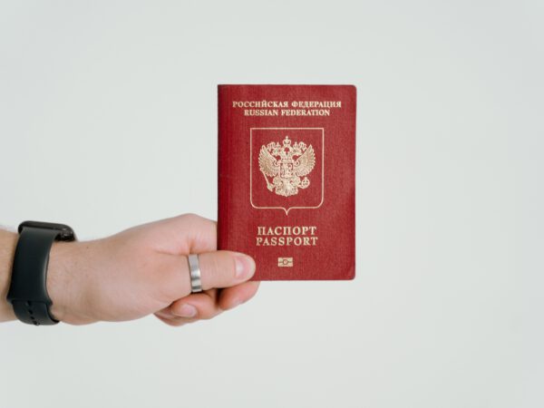 Dłoń z czarną opaską na ręce, trzymająca czerwony paszport na tle białej ściany
